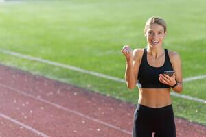 souriant athlétique femme repos après une courir, en portant sa téléphone et sélection la musique à le stade Piste sur une ensoleillé journée. photo