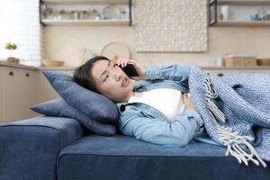 Jeune asiatique femme mensonge malade sur canapé à maison. appels sur une mobile téléphone à le urgence chambre, à le médecin, à le hôpital. couvert avec une couverture, en portant sa estomac, elle se sent mauvais. photo