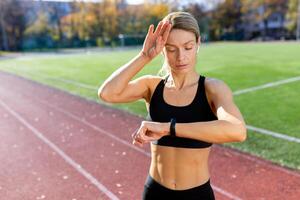 fatigué et dérangé mature femelle athlète vérification cœur taux sur intelligent regarder aptitude bracelet, coureur dans stade après actif exercice et le jogging sur ensoleillé jour, blond femme dans tenue de sport. photo