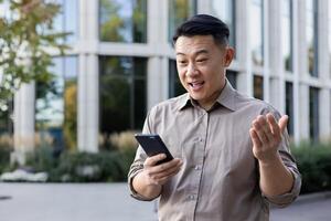 une Jeune asiatique homme est permanent à l'extérieur un Bureau lieu de travail et en portant une téléphone dans le sien main, à la recherche à le écran dans surprise et diffusion le sien mains. photo