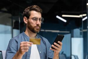 sérieux en pensant homme à l'intérieur Bureau en portant banque crédit carte et téléphone intelligent, homme d'affaire fabrication argent transfert, réservation et achat dans en ligne magasin, réussi entrepreneur. photo