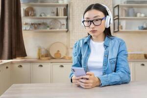 Jeune magnifique concentré asiatique femme écoute à livre audio, podcast. séance à Accueil dans le cuisine portant blanc écouteurs et en portant le téléphone. photo