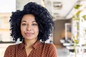 portrait de une Jeune sérieux Afro-américain femme qui travaux dans une restauration établissement, restaurant. permanent dans un tablier à l'intérieur, à la recherche à le caméra. photo