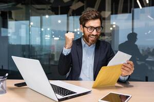 mature homme d'affaire content avec bien nouvelles reçu lettre dans enveloppe, Sénior homme avec barbe en train de lire et souriant travail à l'intérieur Bureau à travail en utilisant ordinateur portable, investisseur célébrer réussite la victoire. photo