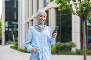 une jeune, de bonne humeur musulman femme portant une hijab, en portant une téléphone intelligent, des promenades en toute confiance à côté de un Bureau bâtiment. photo
