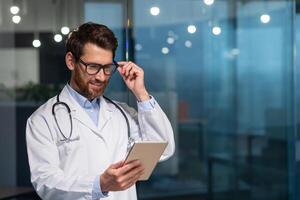 de bonne humeur et souriant Sénior médecin dans des lunettes et médical robe en train de lire message de tablette ordinateur, homme travail à l'intérieur moderne clinique photo