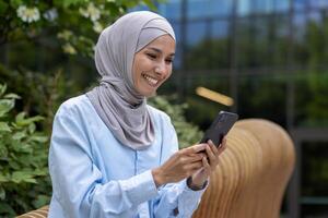 Jeune magnifique musulman femme dans hijab les usages téléphone à l'extérieur Bureau bâtiment, souriant femme lit en ligne nouvelles, parcourt l'Internet pages et cadrans message appel. photo