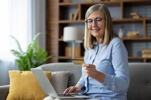 mature femme avec des lunettes sourit comme elle les usages une portable à faire un en ligne achat en utilisant une crédit carte dans une confortable Accueil paramètre. photo