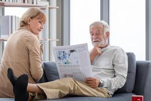 Sénior couple dépenses temps ensemble dans le vivant chambre, retraité homme et femme en train de lire journal, content famille concepts photo