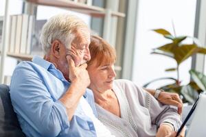 Sénior couple dépenses temps ensemble dans le vivant chambre, femme et homme en utilisant numérique tablette sur le confortable canapé à Accueil photo