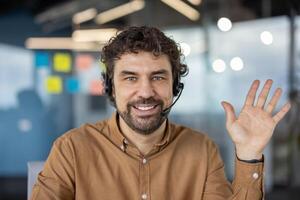 portrait de une de bonne humeur Masculin client un service représentant portant une casque et agitant dans une moderne Bureau réglage photo