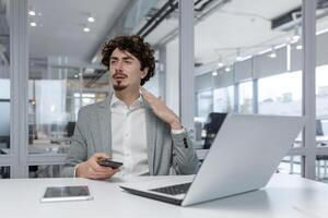 concentré Jeune adulte homme d'affaire avec frisé cheveux travail à le sien bureau dans une moderne Bureau environnement, en utilisant portable et téléphone intelligent. photo