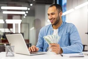 de bonne humeur Masculin entrepreneur en portant en espèces dans main, souriant avec la satisfaction tandis que à la recherche à le sien portable dans une moderne Bureau lieu de travail. photo