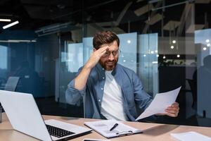 dérangé homme d'affaire derrière papier travail à l'intérieur moderne bureau, mature homme avec barbe en train de lire financier rapports et Compte les documents malheureux avec résultats et déçu avec réalisations. photo