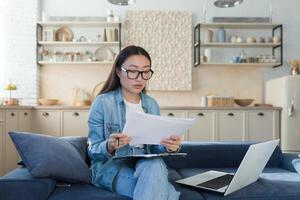 Jeune magnifique Créatif femme asiatique écrivain, journaliste. il est assis à Accueil sur le canapé avec une portable et écrit une livre, un essai, une scénario, un article. détient papier et stylo. photo