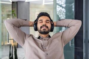 portrait de détendu Indien homme dans écouteurs penché sur retour de chaise avec fermé yeux sur flou Contexte. calme directeur repos dans Bureau avec mains derrière tête et écoute à apaisant playlist. photo