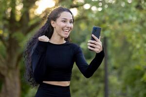 joyeux hispanique femme en plein air, gagnant une défi sur sa téléphone, pompes poing dans excitation, souriant largement. photo