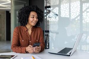 Jeune africain américain femme séance dans le Bureau à le table avec une ordinateur portable, parlant sur une appel et en utilisant le téléphone tandis que souriant. photo