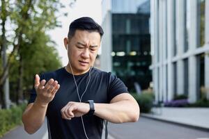 fermer photo. une Jeune asiatique homme Est-ce que des sports, court sur une ville rue dans écouteurs, regards Malheureusement à le résultat sur une intelligent montre, jette le sien mains dans frustration. photo