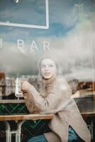 femme dans café regarder en dehors la fenêtre. une femme est assis à une café tableau, habillé dans une confortable chandail et jeans, perdu dans pensée comme elle regards en dehors le fenêtre, capturer une moment de contemplation et relaxation. photo