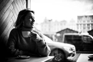 femme profiter café par fenêtre dans ville café. une contenu femme est assis à une café tableau, souriant et regarder en dehors le fenêtre avec une tasse de café dans main. le Urbain scène à l'extérieur Caractéristiques une garé auto. photo