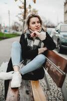 souriant femme dans Urbain paramètre. de bonne humeur femme assise sur banc dans ville, orné dans noir manteau et à carreaux foulard, sourit à caméra. Urbain mode de vie concept avec garé voitures dans Contexte. photo