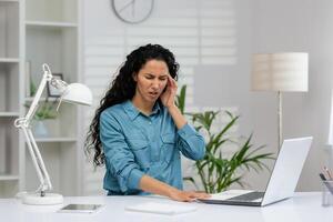 une affligé hispanique femme est en portant sa oreille dans douleur tandis que travail à sa Accueil bureau. elle regards concerné et inconfortable. photo