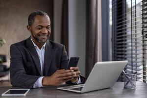 sur de soi africain américain homme d'affaire dans une costume souriant tandis que en utilisant le sien téléphone intelligent et portable dans une moderne Bureau paramètre. photo