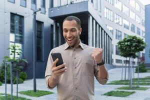 réussi africain américain homme d'affaire à l'extérieur Bureau bâtiment en utilisant téléphone intelligent, souriant et content célébrer la victoire en ligne Jeux, eu bien nouvelles. photo