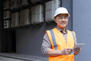 portrait de une souriant Jeune Masculin construction ouvrier dans une difficile chapeau et gilet en portant une tablette dans le sien mains et à la recherche à le caméra. photo