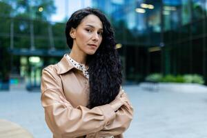 élégant femme d'affaires avec frisé cheveux traversée bras pendant une travail Pause à l'extérieur une entreprise Bureau bâtiment, à la recherche composé. photo