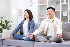 Jeune asiatique Jeune famille, couple, homme et femme dépenser temps ensemble, repos, faire yoga. elles ou ils sont séance sur le canapé dans le lotus position avec leur yeux fermé, souriant, relaxant. photo