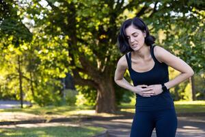 une Jeune femme est permanent dans le parc en plein air, courbé plus de et en portant sa estomac, sentiment sévère douleur après fonctionnement et Faire des sports. photo