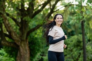 Jeune magnifique femme fonctionnement dans le soir dans le parc, hispanique femme avec frisé cheveux souriant avec contentement, actif mode de vie photo