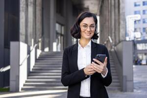 sur de soi femelle professionnel dans affaires tenue souriant tandis que en utilisant une mobile téléphone sur une ville rue avec escaliers dans le Contexte. photo