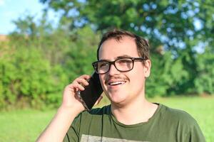 Jeune homme avec lunettes et vert T-shirt parlant sur cellule téléphone à le parc photo