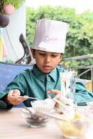 mignonne Indien chef garçon en train de préparer sundae plat comme une partie de non Feu cuisine lequel comprend vanille la glace crème, lutin, coco poudre, fraîchement haché des fruits et fraise sirop. peu enfant en train de préparer nourriture photo