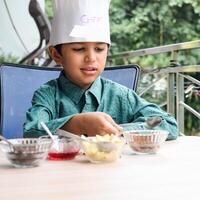 mignonne Indien chef garçon en train de préparer sundae plat comme une partie de non Feu cuisine lequel comprend vanille la glace crème, lutin, coco poudre, fraîchement haché des fruits et fraise sirop. peu enfant en train de préparer nourriture photo