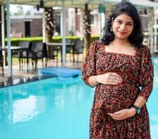 une Enceinte Indien Dame pose pour Extérieur grossesse tirer et mains sur ventre, Indien Enceinte femme met sa main sur sa estomac avec une maternité robe à société parc, Enceinte à l'extérieur maternité tirer photo