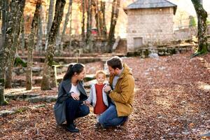 souriant papa et maman sont squat près une peu fille parmi le déchue feuilles dans le forêt photo