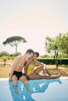 souriant homme baisers femme sur le épaule étreindre sa taille de derrière tandis que séance sur le bord de le bassin photo