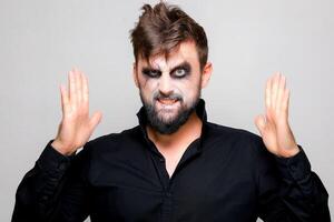 une homme avec une barbe et Mort-vivant maquillage sur Halloween détient le sien mains dans de face de lui photo