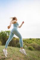 bas vue de un athlétique fille le jogging dans le parc. magnifique blond caucasien femme dans bleu serré survêtement. blond fille à un Extérieur formation session photo