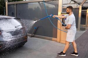 une homme détient une mousse pulvérisateur de lequel mousse mouches en dehors sur une auto. une voiture à une soi un service voiture laver. photo