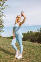 fille prise selfies dans le parc en portant sa téléphone dans de face de son. magnifique blond caucasien femme dans bleu serré survêtement. blond fille à un Extérieur formation session photo