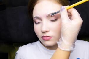 une Jeune blond modèle dans le sourcil laminage procédure, le Maître les usages une brosse à appliquer stratification composés à le les sourcils photo