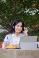 Bureau magnifique asiatique femme dans Extérieur café. gâté souriant fille écoute à la musique tandis que dactylographie Faire travail dans de face de le portable à la recherche à le caméra pour contenu ou promotions photo