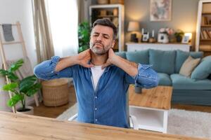 homme d'affaire sentiment douleur dans cou après séance à le table avec portable. fatigué homme Souffrance de Bureau syndrome car de longue heures ordinateur travail. il est masser le sien tendu cou muscles photo