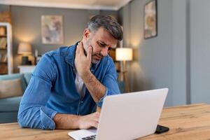 homme d'affaire sentiment douleur dans cou après séance à le table avec portable. fatigué homme Souffrance de Bureau syndrome car de longue heures ordinateur travail. il est masser le sien tendu cou muscles photo