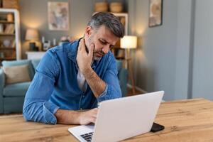 homme d'affaire sentiment douleur dans cou après séance à le table avec portable. fatigué homme Souffrance de Bureau syndrome car de longue heures ordinateur travail. il est masser le sien tendu cou muscles photo